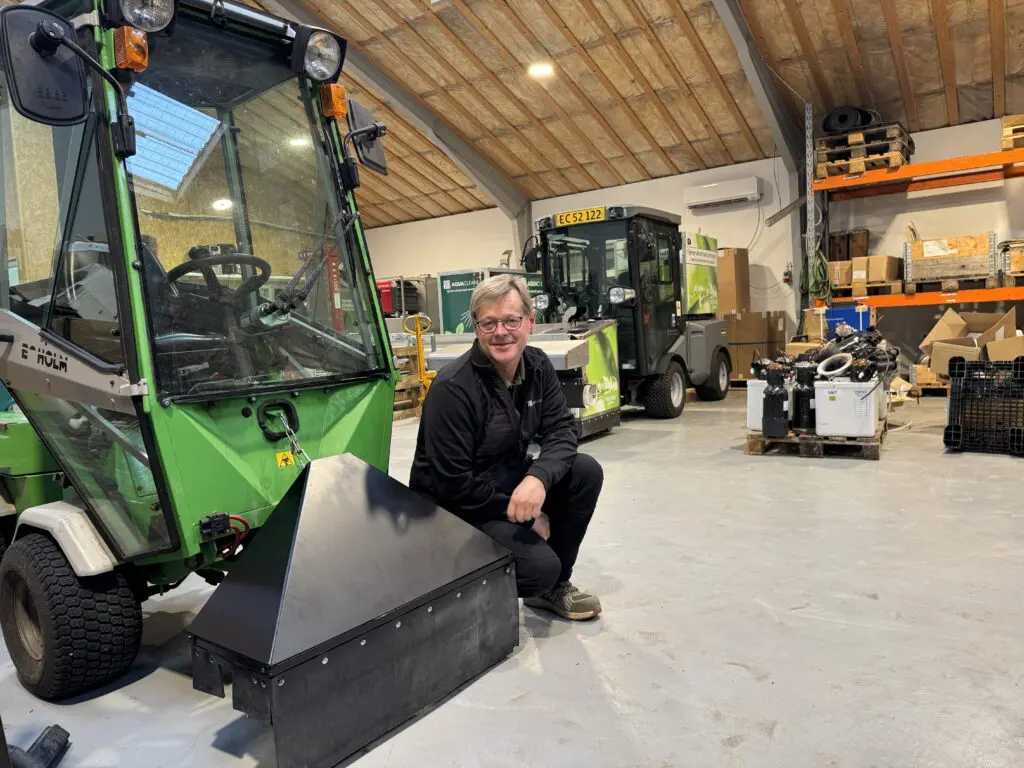 En smilende mand fra Aqua Cleaner ved en grøn Greenheat ukrudtsmaskine i et værksted.