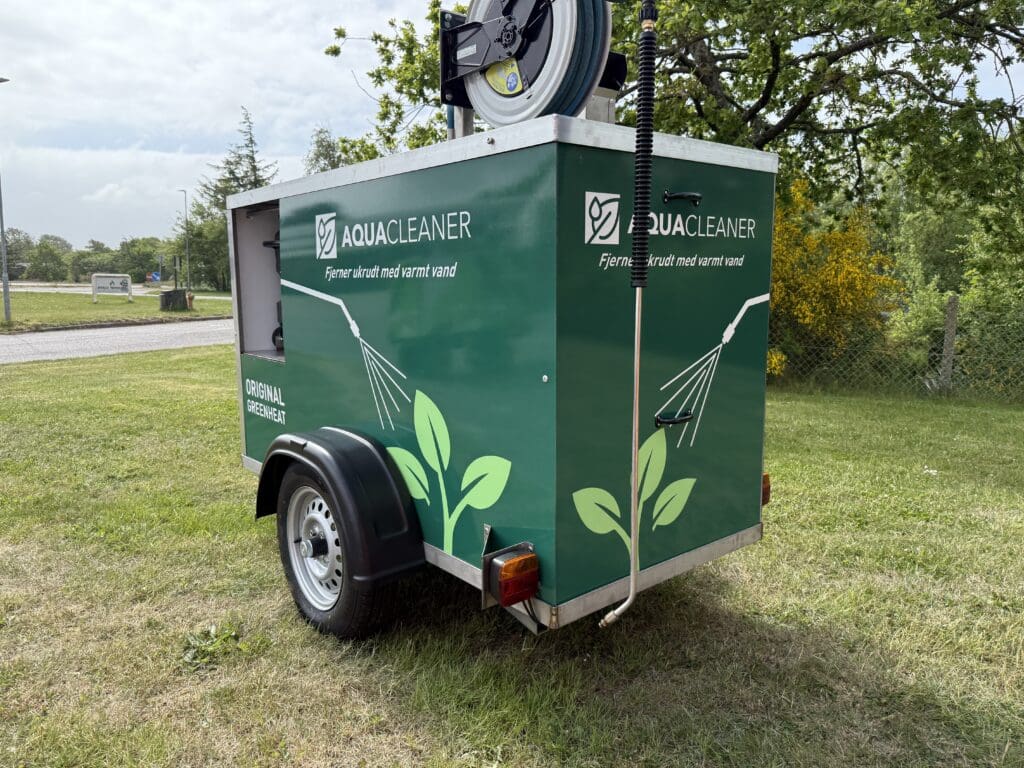 A green Aqua Cleaner trailer for environmentally friendly weed control using 105-degree hot water, parked on a grassy area.