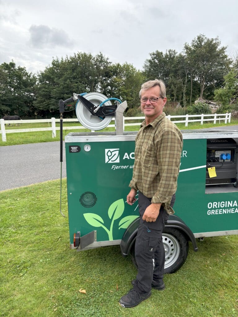 En mand står ved siden af en grøn Aqua Cleaner Greenheat trailer til miljørigtig ukrudtsbekæmpelse med 105 grader varmt vand.