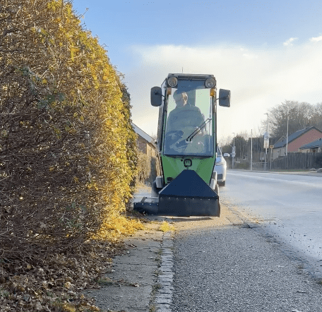 Grøn Aqua Cleaner maskine med fører fejer blade og snavs fra fortov langs tæt hæk på solrig dag.