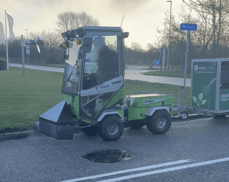 Grøn og hvid Holm Z180+ maskine med fører sprøjter vand på vej, trækker Aqua-Farm trailer til ukrudtsbekæmpelse.