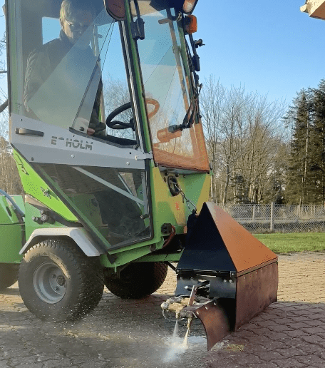 Mand i grøn E-HOLM maskine sprøjter varmt vand fra en dyssebom på fliser til effektiv ukrudtsbekæmpelse.