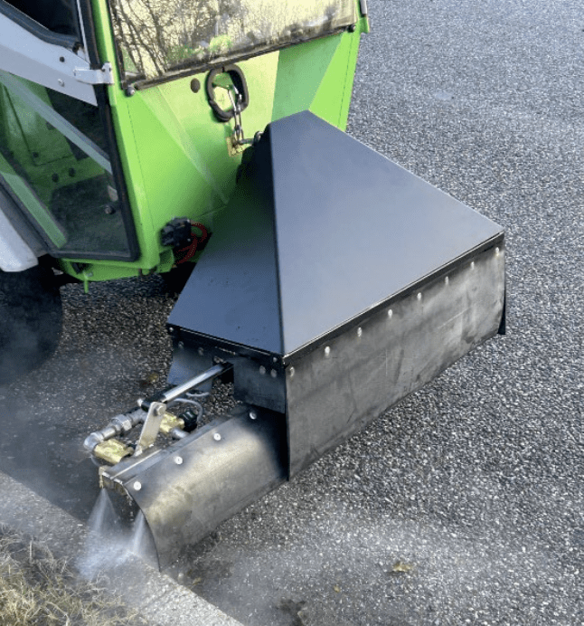 Grøn Aqua Cleaner maskine med frontmonteret dyssebom, der sprøjter vandstråler på ukrudt langs en vejrabat for effektiv bekæmpelse.