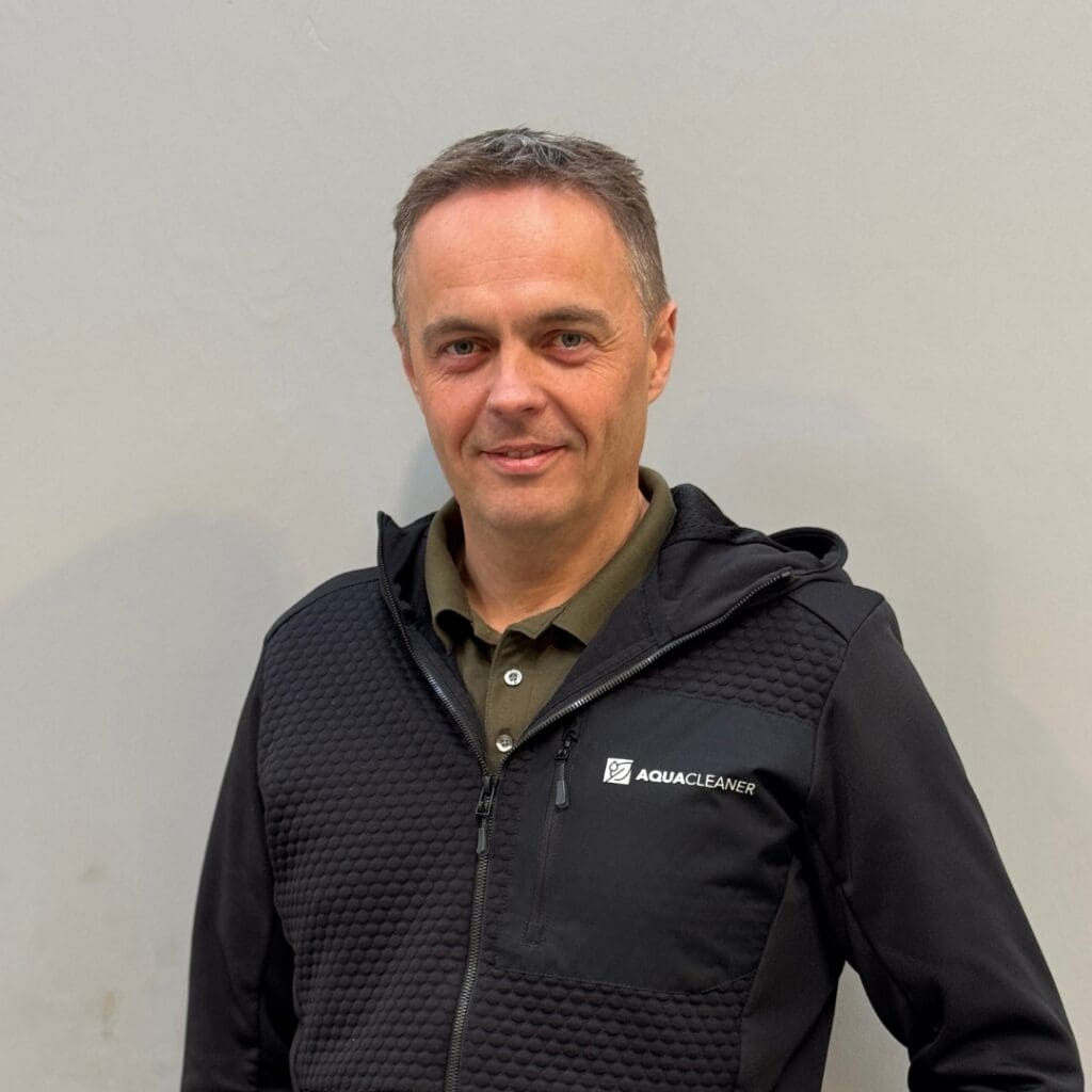Tommy, ein lächelnder Mann mit kurzen grauen Haaren, trägt eine schwarze Aqua-Cleaner-Jacke und ein grünes Polohemd. Er ist Experte für Hochdruckreiniger und Unkrautbekämpfung.